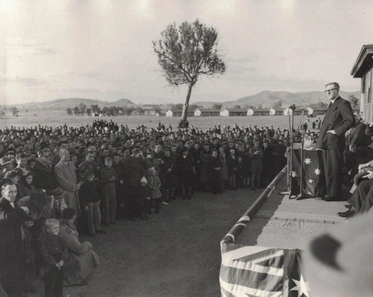 Arthur Calwell stands on a platform with microphones, addressing a crowd of people. In the distance is a row of long buildings. Arthur Calwell stands on a platform with microphones, addressing a crowd of people. In the distance is a row of long buildings.