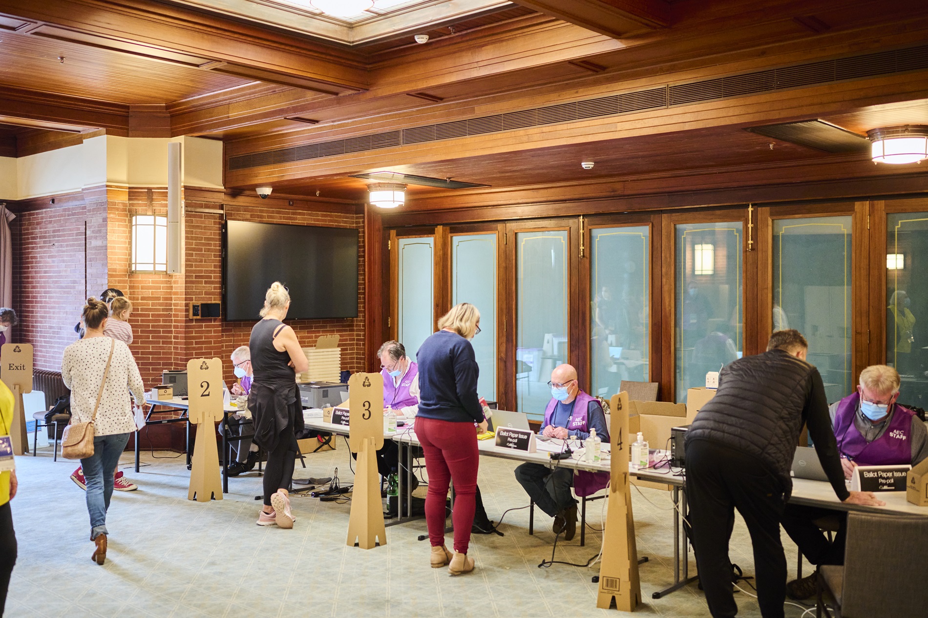 Four Australian Electorial Commission staff sit in a row at a long table wearing face masks and purple vests, and people are lined up, one in front of each staff member, to collect their ballots. Four Australian Electorial Commission staff sit in a row at a long table wearing face masks and purple vests, and people are lined up, one in front of each staff member, to collect their ballots.