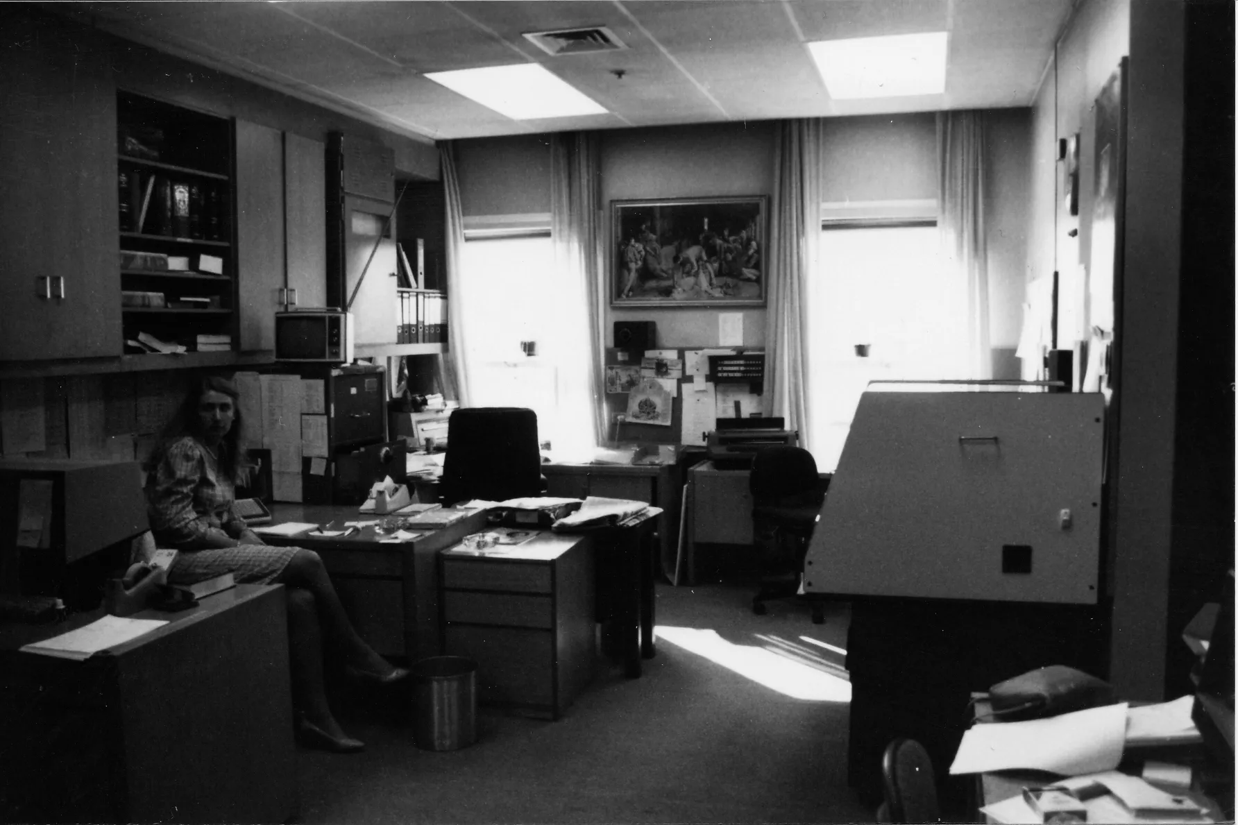 The black and white photograph shows Heather Mittag dressed in a plaid skirt suit sitting on a chair in a small open plan office. Several desks are dominated by large typewriters and computer monitors. The walls are filled with pinboards containing notes and reference sheets and there are numerous filing cabinets hold. On the back wall is a framed print of the painting ‘The Shearing of the Rams’ by Tom Roberts. The black and white photograph shows Heather Mittag dressed in a plaid skirt suit sitting on a chair in a small open plan office. Several desks are dominated by large typewriters and computer monitors. The walls are filled with pinboards containing notes and reference sheets and there are numerous filing cabinets hold. On the back wall is a framed print of the painting ‘The Shearing of the Rams’ by Tom Roberts.