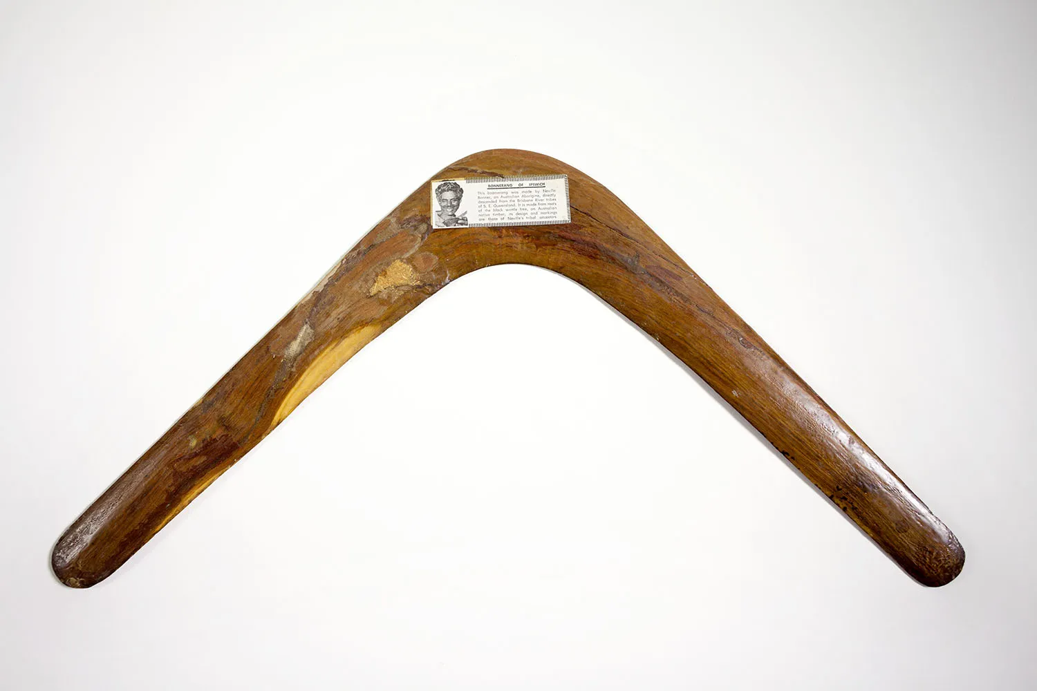 A wooden boomerang with a white descriptive label featuring text and a man's face. A wooden boomerang with a white descriptive label featuring text and a man's face.