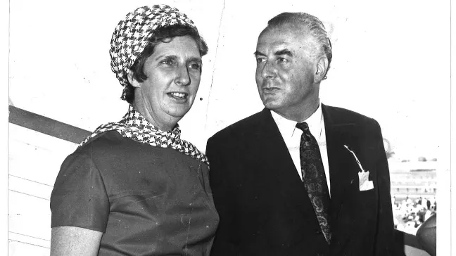 A black and white photograph of Margaret and Gough Whitlam. Mr Whitlam wears a suit and tie while Mrs Whitlam wears a check scarf on her head and neck.