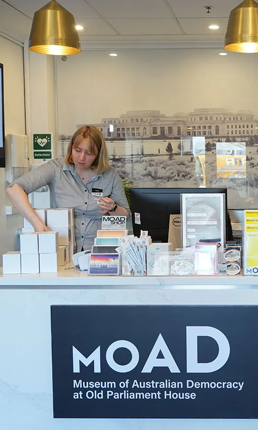 A museum staff member stands behind the shop counter at the Museum of Australian Democracy at Old Parliament House wearing a black and white check shirt and a name badge. A museum staff member stands behind the shop counter at the Museum of Australian Democracy at Old Parliament House wearing a black and white check shirt and a name badge.