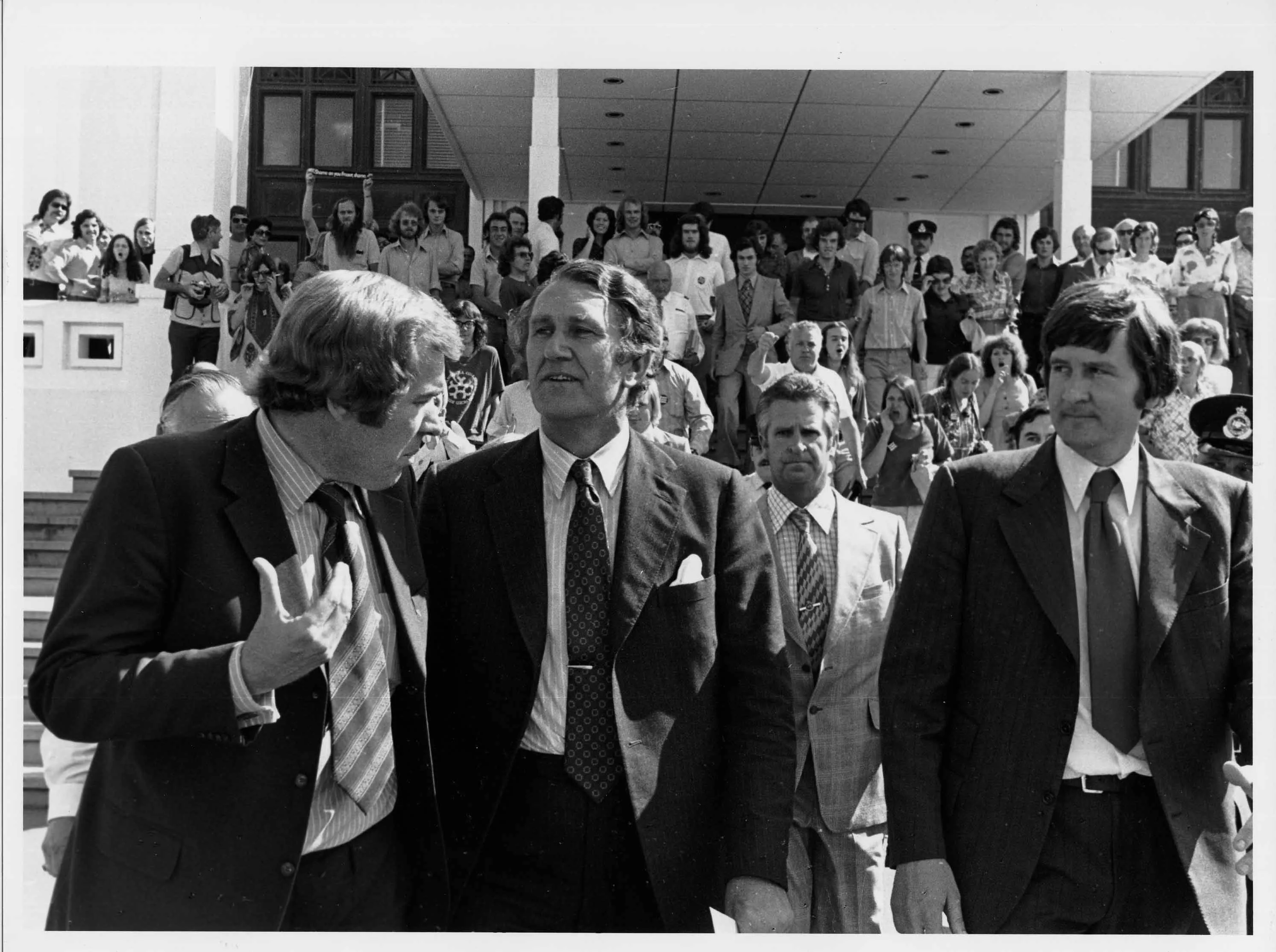 Malcolm Fraser walks away from Parliament House with two other men in suits. A crowd of people watch from the steps behind them.  