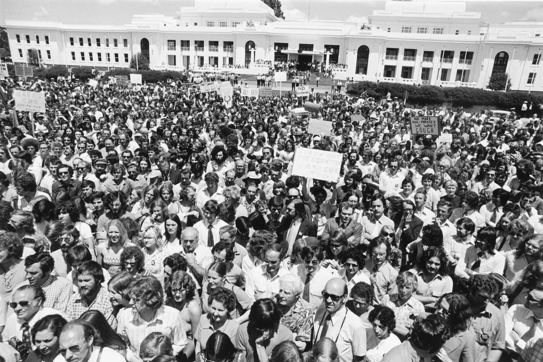 A large crowd of people gathered outside Parliament House, some holding placards. 
