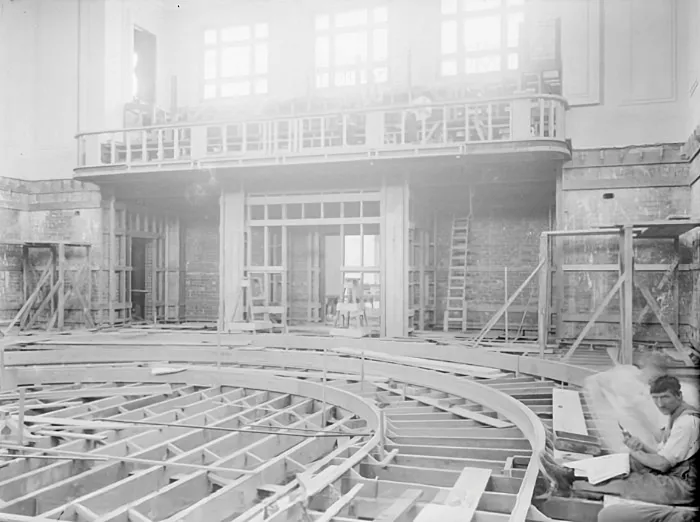 This black and white photograph depicts the House of Representatives Chamber partway through construction. The horseshoe shape of the chamber seating is visible in the timber framework. The public gallery is partly constructed and westerly light pours through the high windows. There are timber frames and a ladder against the back wall of the chamber. In the right foreground, a lone construction worker sits between the timber floor beams, reading a newspaper and smoking a pipe. This black and white photograph depicts the House of Representatives Chamber partway through construction. The horseshoe shape of the chamber seating is visible in the timber framework. The public gallery is partly constructed and westerly light pours through the high windows. There are timber frames and a ladder against the back wall of the chamber. In the right foreground, a lone construction worker sits between the timber floor beams, reading a newspaper and smoking a pipe.